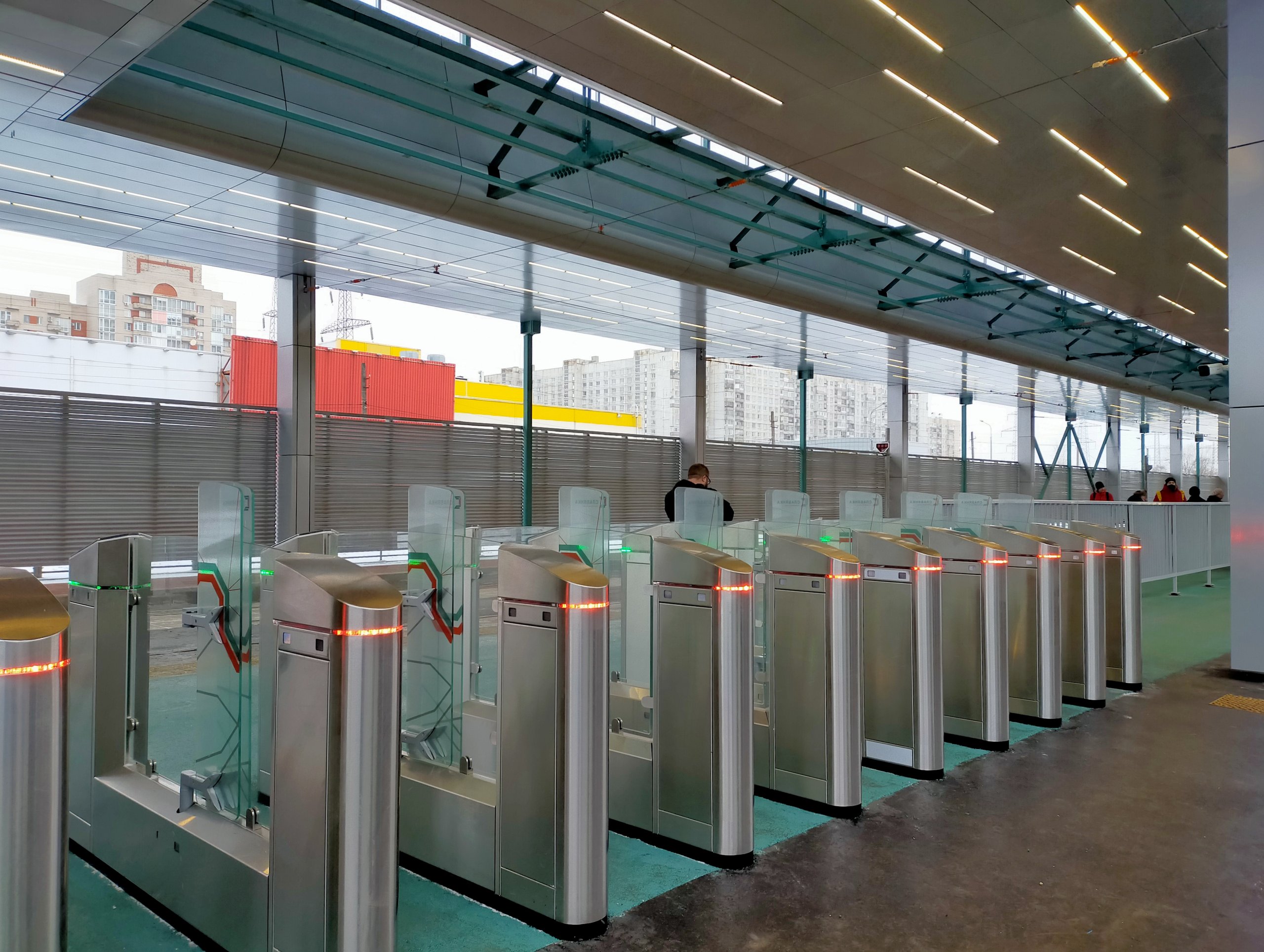 A row of stainless steel ticket gate turnstiles with glass flaps and orange indicator lights at a covered tram station
